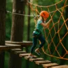 Dziewczynka w uprzęży pokonuje trasę linową w lesie – zbliżenie, styl tilt-shift, lina pomarańczowa, drewniane platformy