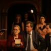 Scena teatralna z aktorami w trakcie śledztwa – kobieta trzymająca szkatułkę z naszyjnikiem, obok niej policjant i podejrzani, czerwone kurtyny i stare wnętrze teatru