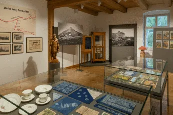 Widok wystawy w muzeum etnograficznym, z porcelaną, materiałami blaudruk, mapą Śląskiej Kolei Górskiej i drewnianą rzeźbą w sali oświetlonej światłem dziennym