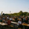 Tłum uczestników festiwalu piwa siedzących przy drewnianych ławach na świeżym powietrzu, z widokiem na stoiska browarów i zielone drzewa w tle. Zdjęcie wykonane z lewego boku placu, w poziomie, w świetle zachodzącego słońca.