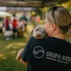 Kobieta w koszulce GRUPY RATUJ przytulająca szczeniaka podczas pikniku charytatywnego w Parku Grabiszyńskim we Wrocławiu, w tle rozmyta grupa uczestników wydarzenia na świeżym powietrzu.