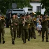 Wojskowa orkiestra maszerująca przez trawiaste pole podczas rodzinnego pikniku militarnego REKON we Wrocławiu, z pojazdem opancerzonym w tle i tłumem widzów po lewej stronie