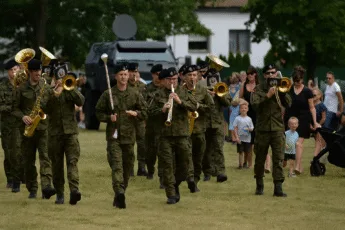 Wojskowa orkiestra maszerująca przez trawiaste pole podczas rodzinnego pikniku militarnego REKON we Wrocławiu, z pojazdem opancerzonym w tle i tłumem widzów po lewej stronie